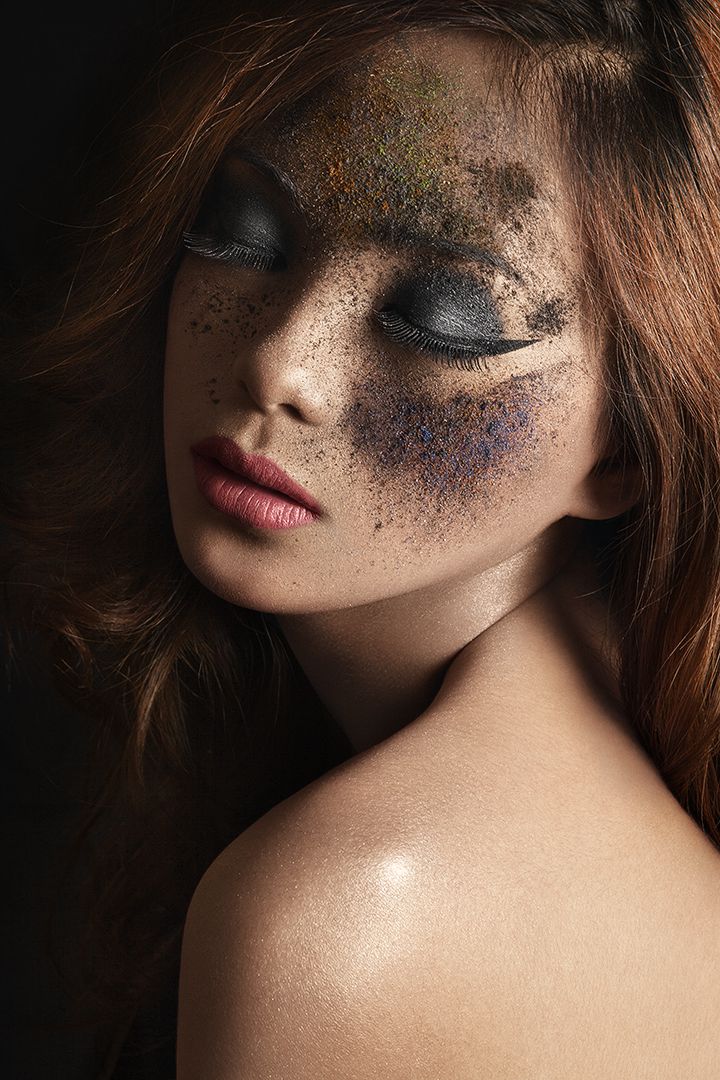 Artistic beauty close-up with scattered pigment powder across the face, eyes closed, moody dark lighting