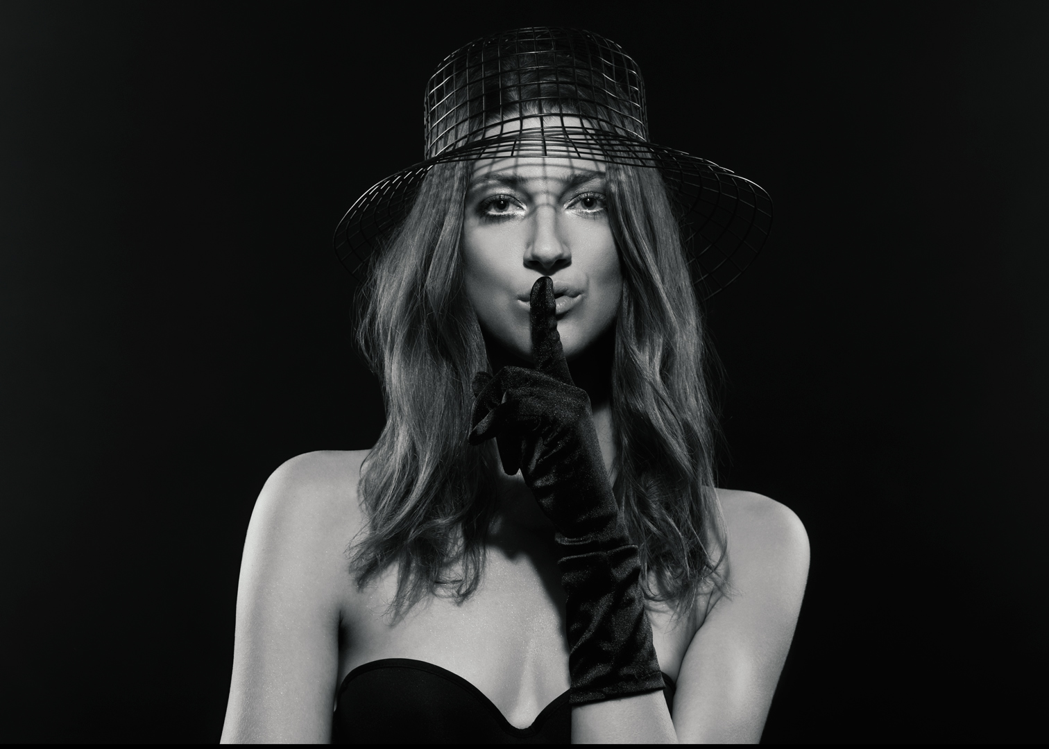 Black and white editorial portrait with wire cage hat and satin gloves in a hushing gesture